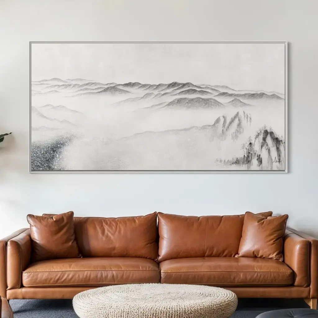 A wide panoramic landscape artwork depicting foggy mountains, hanging above a brown leather sofa in a living room.